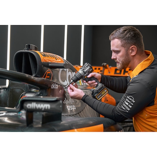 A person using a DEWALT cordless power drill on the body of an orange and black race car. The person's face is blurred.