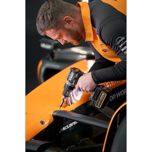 A person with their face blurred is using a DEWALT power drill (DCD85MP2T) on an orange racing car, wearing an orange and black uniform.