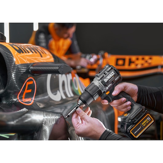 A person using a DEWALT cordless power drill (DCD85MP2T) to work on the exterior of a carbon fiber race car. The car has black and orange detailing, and another person in an orange uniform is blurred in the background.