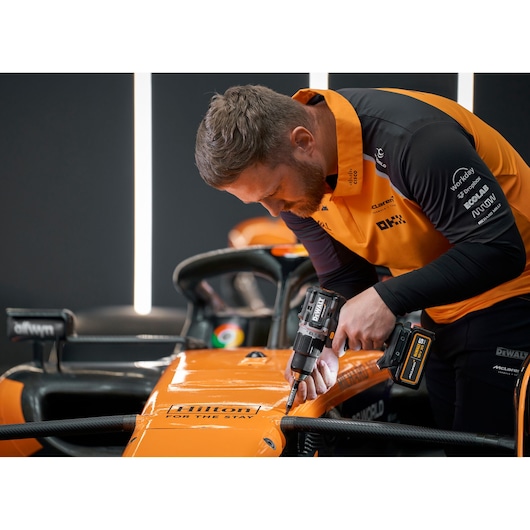 A person in an orange and black team uniform uses a DEWALT cordless drill to work on the body of an orange race car, with the car branded 'Hilton' and surrounded by modern lighting.