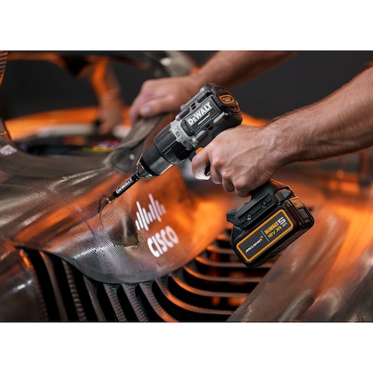 A close-up of a person's hands using a DEWALT cordless drill on the surface of a carbon fiber component, featuring Cisco branding and vibrant orange lighting.