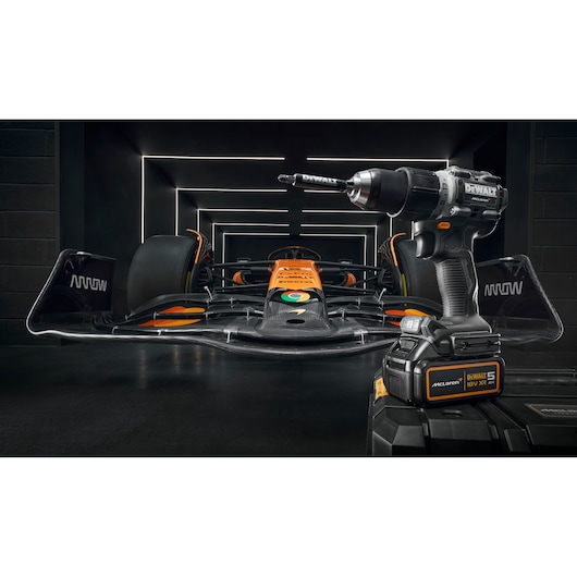 A DEWALT cordless power drill is displayed prominently in the foreground, with a black and orange Formula 1 race car positioned behind it in a modern, illuminated garage setting.