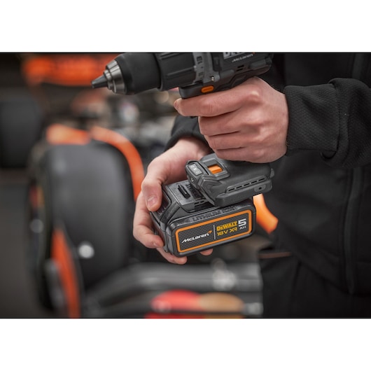 Close-up of a person holding a DEWALT cordless drill with an 18V XR 5.0Ah McLaren battery, with a blurred orange and black race car in the background.