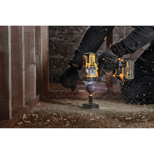A person using a DEWALT cordless power drill to drill a hole in a wooden surface. Wood shavings are flying around as the drill is in use.
