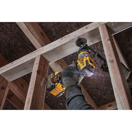 A person using a DEWALT cordless drill to bore a hole through a wooden beam in a construction setting. The user is wearing gloves and sawdust is visible as the drill operates.