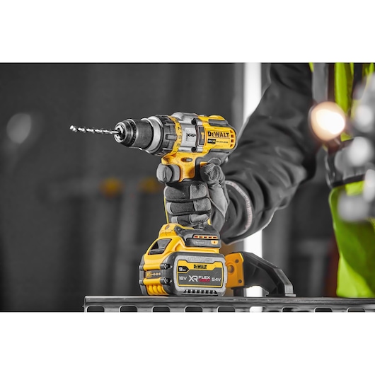 A close-up of a person wearing work gloves using a DEWALT cordless power drill with a brushless motor. The drill is yellow and black and features an XR FLEXVOLT 54V 18V 9.0Ah battery. The background is out of focus, highlighting the drill and the user's hand.