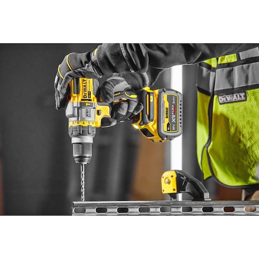 A close-up view of a worker using a DEWALT DCD999X1 cordless drill to drill into a metal beam. The worker is wearing black gloves and a yellow safety vest with a DEWALT logo.