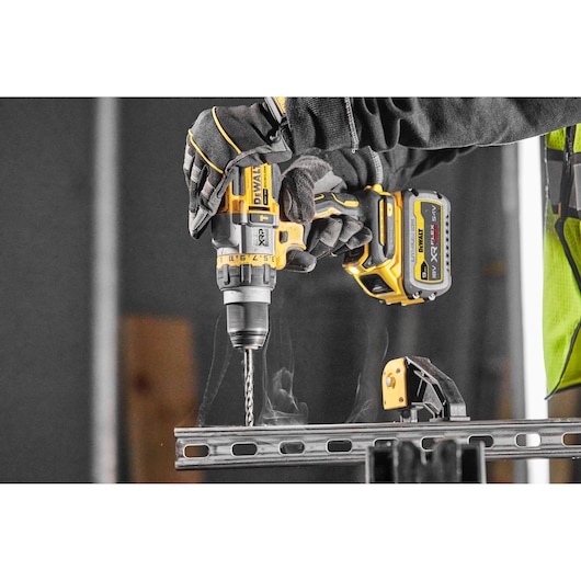 Close-up of a person wearing gloves using a DEWALT DCD999X1 cordless drill to drill through a metal beam, with visible smoke rising from the material.