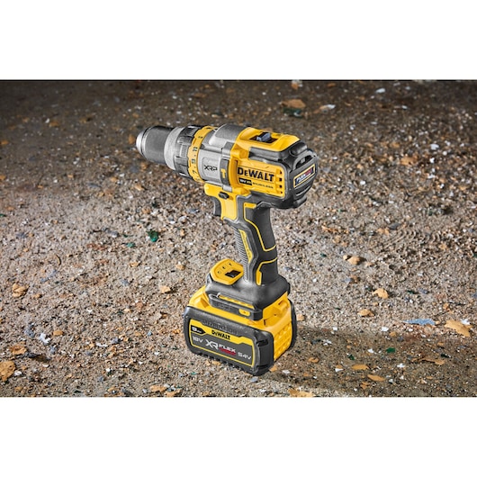 A DEWALT 18V XR FLEXVOLT cordless drill, model DCD999X1, standing upright on a rough, dusty surface. The drill is yellow and black with a large battery attached at the base.