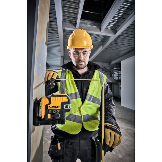 A person wearing a yellow hard hat, high-visibility vest, and work gloves is using a DEWALT DCE0822D1G green laser level tool attached to a metal column in an industrial or construction environment.