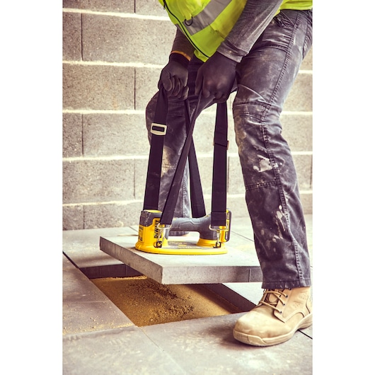 A worker uses a DEWALT DCE590 tool with straps to lift a heavy concrete slab during construction. The worker is wearing gloves, work boots, and a high-visibility vest. The scene shows the slab being positioned over a sand-filled space among concrete blocks.