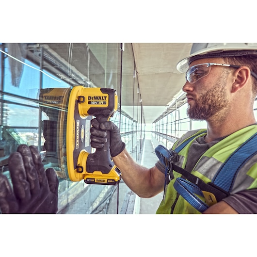 A construction worker wearing safety gear uses a DEWALT 18V XR cordless power tool (SKU: DCE590) to handle or install glass panels on the exterior of a modern building.