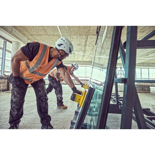Two construction workers wearing safety helmets and orange vests are using DEWALT DCE590 tools to move a large glass pane in an industrial building.