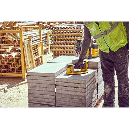 A person wearing a safety vest and work gloves uses a yellow DEWALT power tool on a stack of rectangular concrete slabs at a construction site. Various construction materials and equipment are visible in the background.