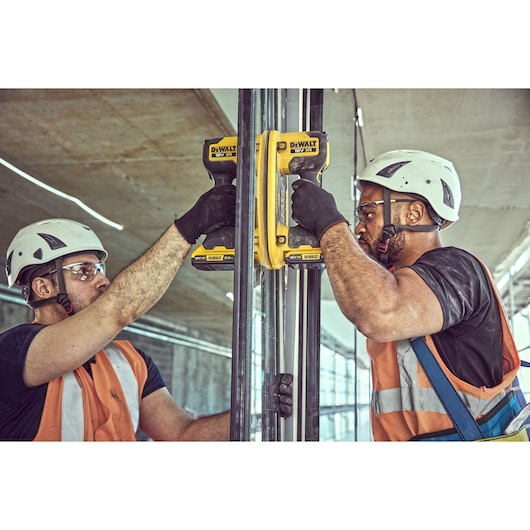 Two workers wearing safety helmets and orange vests use DEWALT DCE590 cordless tools to fasten metal framing at a construction site. Their faces are blurred for privacy.
