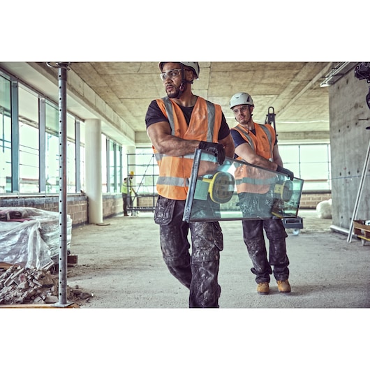 Two construction workers wearing orange safety vests and hard hats carry a large glass panel using a DEWALT suction lifter inside a building under construction.