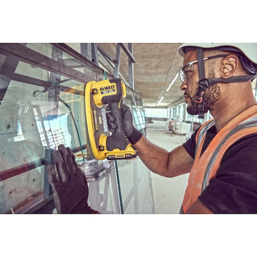 A person wearing gloves and a safety vest uses a DEWALT DCE590 cordless tool to handle a sheet of glass at a construction site. The individual's face is blurred for privacy.