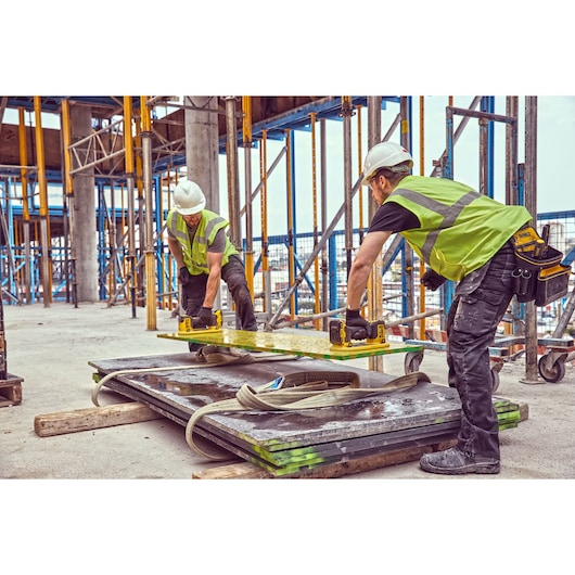 Two construction workers wearing safety vests and helmets are using DEWALT tools to lift a large metal sheet at a construction site with scaffolding and concrete pillars.