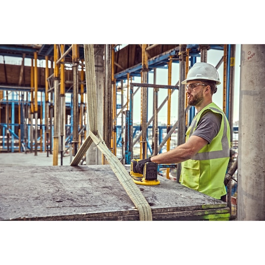 A construction worker wearing a reflective vest and hard hat operates a DEWALT DCE590 tool on a large concrete slab, secured with heavy-duty straps, at a building site with scaffolding and structural columns.