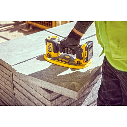 A person wearing work gloves uses a DEWALT DCE590 cordless vacuum lifter to lift a concrete slab. The DEWALT vacuum lifter is yellow and black, positioned on top of stacked concrete slabs at a construction site.