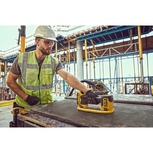 A worker wearing a safety vest, gloves, and a hard hat is using a DEWALT DCE590 tool on a construction site. The tool is placed on a large flat surface and there is scaffolding in the background.