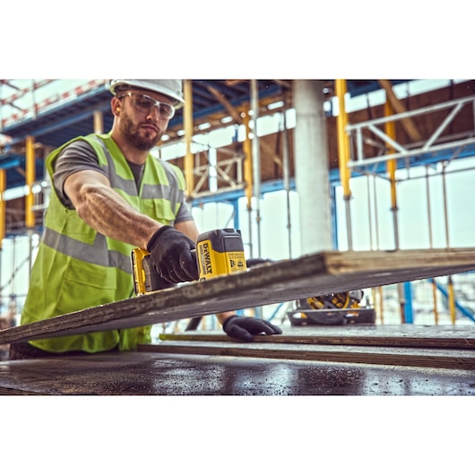 A person wearing a high-visibility vest and gloves is using a DEWALT DCE590 cordless tool to work with a piece of material at a construction site. Scaffolding and building structures are visible in the background.