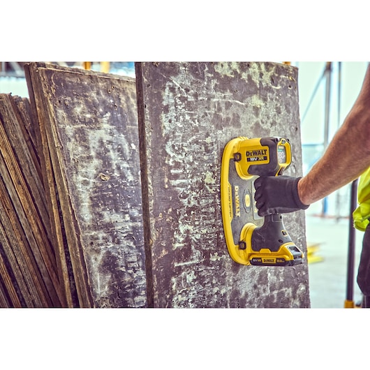 A person wearing a black glove uses a DEWALT 18V XR power tool to sand an old, worn wooden board. Several similar boards are stacked in the background, and the scene appears to be at a construction or renovation site.