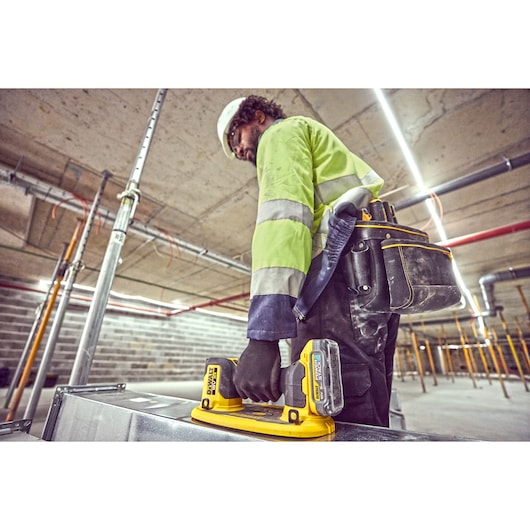 A construction worker wearing a high-visibility jacket and tool belt is using a DEWALT DCE590 tool in an industrial setting with exposed pipes and concrete ceilings.