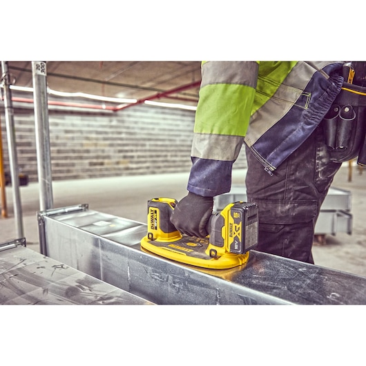 A person wearing work gloves and a high-visibility jacket uses a DEWALT cordless tool on a metal surface in an industrial setting.