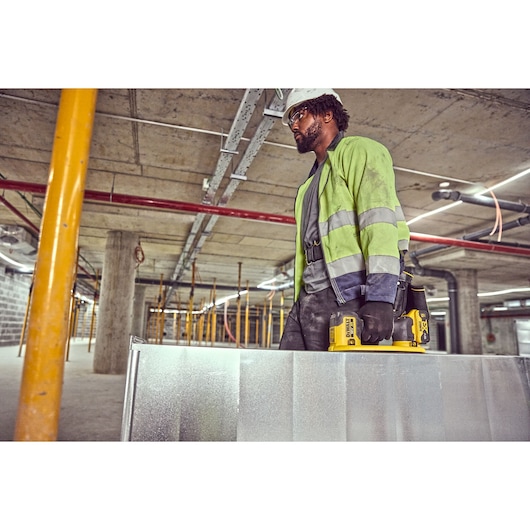 A construction worker wearing a high-visibility jacket and gloves is holding a DEWALT DCE590 tool and carrying a large metal sheet inside an unfinished building with exposed concrete, pipes, and pillars.