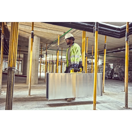 A person wearing a safety helmet and high-visibility jacket carries a large metal case with yellow handles inside a construction site with concrete floors, columns, and support beams.