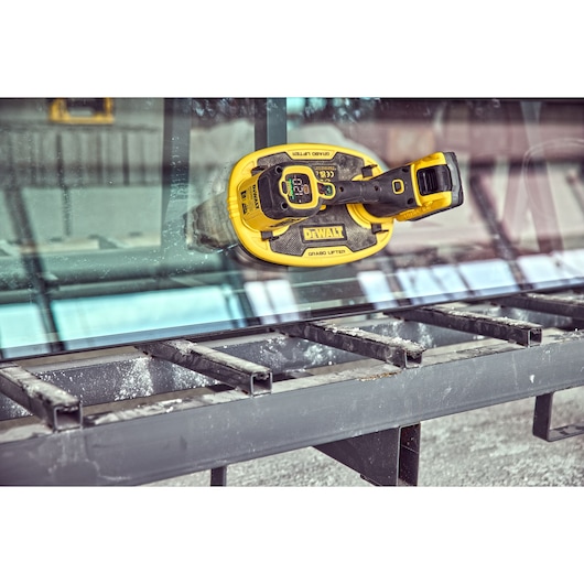 A DEWALT DCE590 battery-powered glass lifter attached to a large glass pane, positioned above a metal frame structure in an industrial environment.