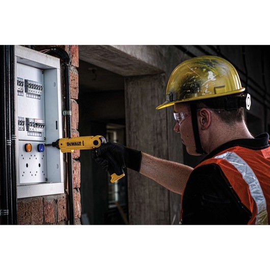 A person wearing a yellow safety helmet and orange reflective vest uses a yellow DEWALT cordless screwdriver (model DCF008) to work on an electrical panel mounted on a brick wall.