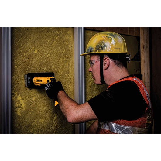 A worker wearing a yellow hard hat, orange safety vest, and black gloves uses a DEWALT DCF008 tool to install or prepare a section in a yellow textured wall on a construction site. The worker's face is blurred for privacy.