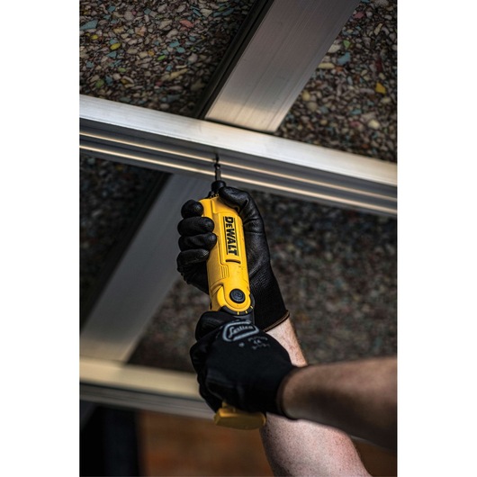 A person wearing black work gloves is using a yellow DEWALT cordless screwdriver to drive a screw into a metal frame, viewed from below.