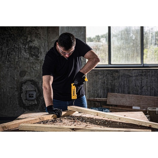 A person wearing black gloves is using a yellow DEWALT cordless drill to work on wooden planks in a workshop with a concrete wall and large window in the background. The person's face is obscured.