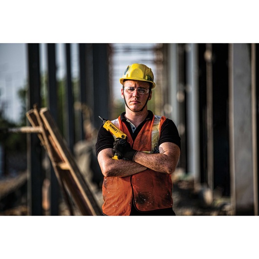 A person wearing a yellow hard hat and orange safety vest stands at a construction site, holding a yellow DEWALT power tool with crossed arms. The face is blurred for privacy.