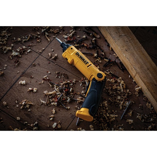 A yellow and black DEWALT cordless screwdriver (SKU: DCF008) lies on a wooden floor, surrounded by wood shavings and screws, with a piece of lumber nearby.