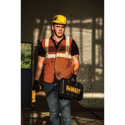 A construction worker wearing a yellow hard hat, orange safety vest, and gloves stands indoors holding a DEWALT tool case in one hand and a yellow DEWALT power tool in the other hand.