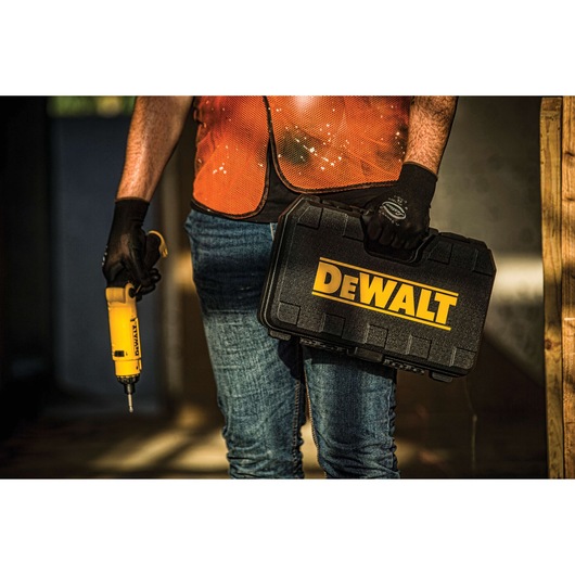 A person wearing an orange safety vest and gloves holds a yellow DEWALT power tool in one hand and a black DEWALT carrying case in the other hand.