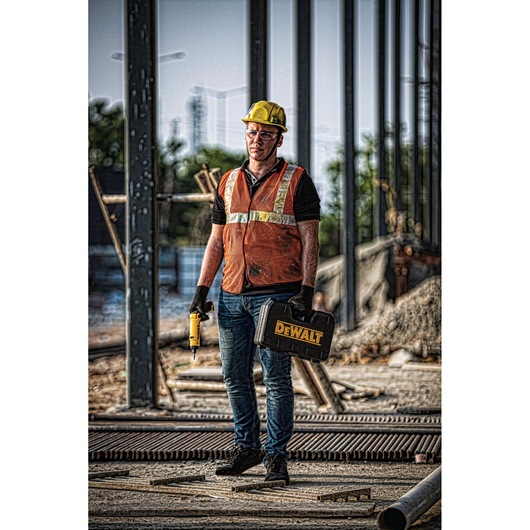 A construction worker wearing a yellow hard hat, orange safety vest, and gloves stands on a building site holding a DEWALT toolbox and a yellow cordless drill. The worker's face is blurred, and steel frameworks are visible in the background.