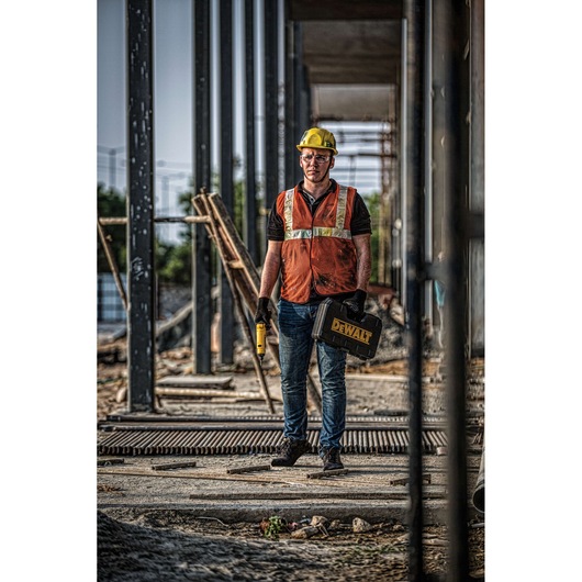 A construction worker wearing a yellow hard hat and an orange safety vest stands at a building site holding a DEWALT tool case and a cordless power tool. The worker is surrounded by steel structures and construction materials.