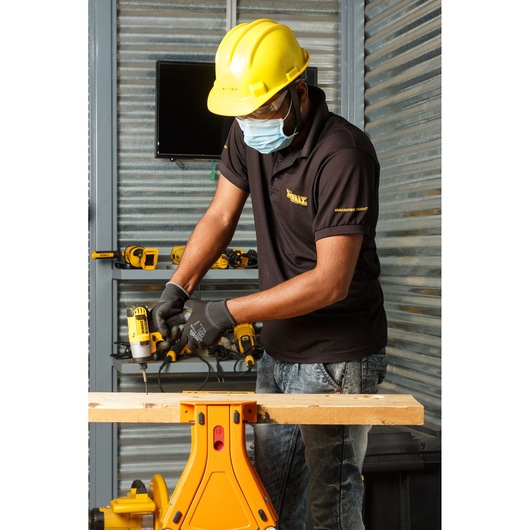 A person wearing a yellow hard hat and black DEWALT shirt uses a DEWALT DCF815D2 power tool to drill into a piece of wood, which is secured on a yellow work stand. The scene is set in a workshop with DEWALT tools visible on shelves in the background.