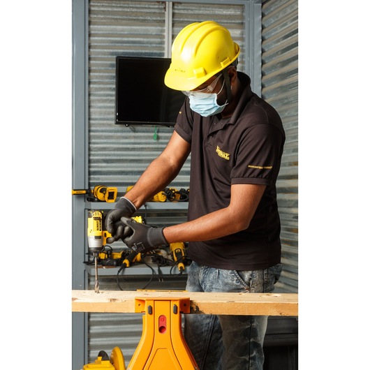 A person wearing a yellow hard hat and black DEWALT shirt is drilling into a wooden plank using a yellow DEWALT cordless drill. The individual is in a workshop with various DEWALT tools visible on the shelves in the background. The person's face is blurred for privacy.