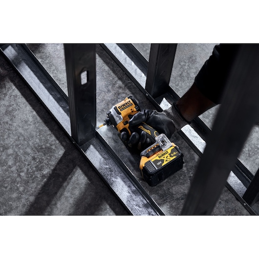 A person wearing a work glove uses a DEWALT cordless impact driver to fasten a screw into a metal frame on a concrete floor.
