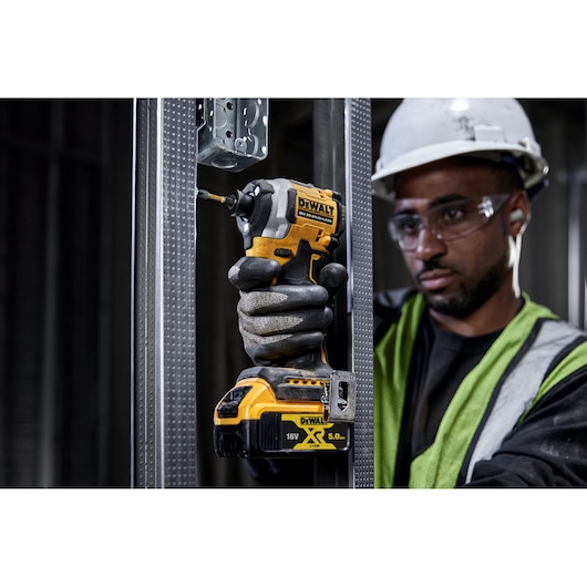A person wearing safety gear is using a DEWALT DCF850N 18V cordless impact driver to drill into a metal frame at a construction site.