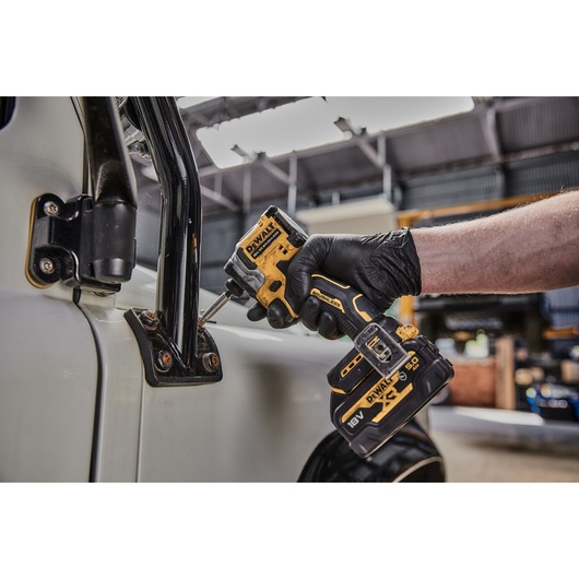A person wearing a black glove is using a DEWALT DCF850N compact impact driver to fasten screws on a vehicle door in an industrial workshop.