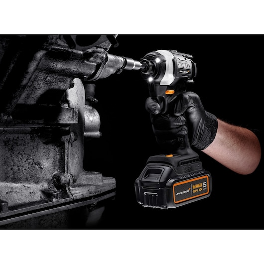 A person wearing a black glove is using a DEWALT McLaren 18V impact driver with a 5Ah battery to work on a metallic