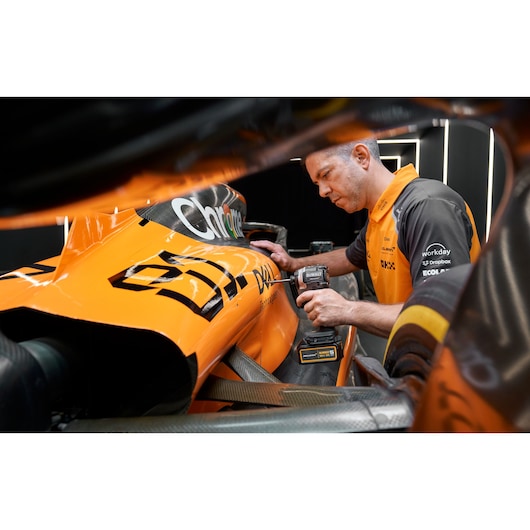 A person wearing an orange and black shirt is using a DEWALT power tool to work on the body of an orange race car. The