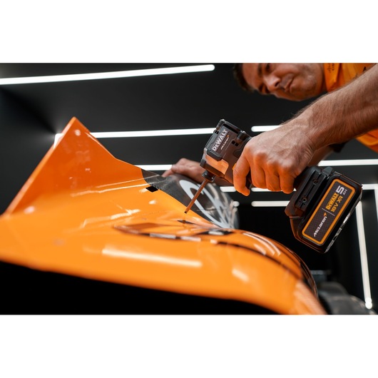 A person uses a DEWALT cordless power tool to drive a screw into the surface of an orange vehicle part, with a modern